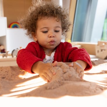 Ein kleines Kind mit lockigem Haar und rotem Pullover spielt drinnen mit Sand, wobei es beide Hände benutzt, um den Sand auf einer flachen Oberfläche zu formen oder zu bewegen. Das natürliche Licht kommt von den großen Fenstern im Hintergrund.