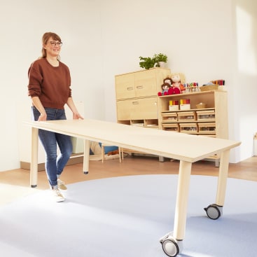 Eine Person mit Brille, braunem Pullover und Jeans bewegt einen hellen Tisch mit Rollen in einem hellen Raum, der mit Regalen, Schränken, Kinderspielzeug und Unterrichtsmaterialien ausgestattet ist.