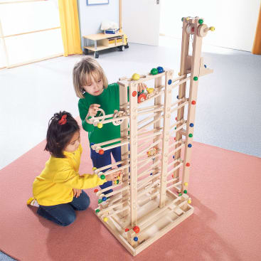 Zwei Kinder sitzen und knien auf einem orangefarbenen Teppich und spielen mit einer großen hölzernen Murmelbahn mit mehreren Rampen und bunten Bällen in einem hellen Raum mit hellen Wänden und Böden.