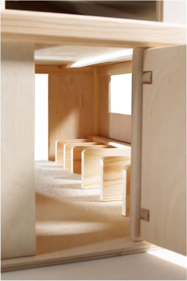 Nahaufnahme eines kleinformatigen Architekturmodells aus Holz, das einen Raum mit vier hölzernen bankähnlichen Strukturen und einem Fenster an der rechten Wand zeigt. Die Tür auf der rechten Seite ist teilweise geöffnet.