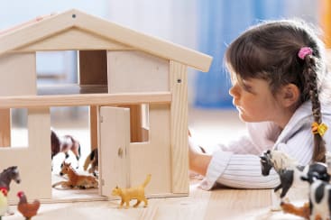 Ein junges Mädchen mit braunem Haar in Zöpfen liegt auf dem Boden neben einer hölzernen Spielzeugscheune und ordnet Plastiktierfiguren, darunter Pferde, Kühe und eine Katze, um die Scheune herum an.