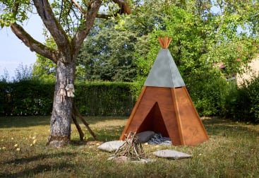Ein hölzernes Spiel-Tipi steht im Gras neben einem Baum, an dessen Stamm ausgestopfte Tiere hängen. Im Inneren des Tipis befinden sich Kissen und vor dem Tipi ein kleines Arrangement aus Stöcken und Steinen. Im Hintergrund sind Sträucher und Bäume zu sehen.