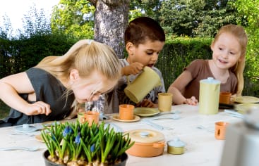 Drei kleine Kinder sitzen an einem weißen Tisch im Freien und spielen mit Spielzeuggeschirr und -tassen eine Teeparty. Ein Kind schenkt aus einer Spielzeug-Teekanne ein, während die anderen zusehen und lächeln. Eine Topfpflanze steht im Vordergrund.