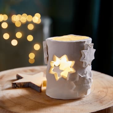 Ein weißer Keramik-Kerzenhalter mit sternförmigen Aussparungen steht auf einer Holzfläche. Von innen leuchtet warmes Licht, und im verschwommenen Hintergrund sind gelbe Lichterketten zu sehen. Ein Holzstern liegt neben dem Halter.