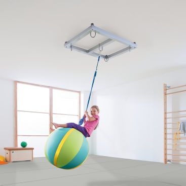 Ein kleines Kind in einem lila Outfit sitzt auf einem großen bunten Therapieball, der an einem blauen Seil in einem hellen Raum mit Turngeräten von der Decke hängt.