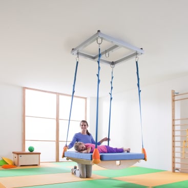 Eine Frau kniet neben einem Kind, das auf einer großen, rechteckigen Plattformschaukel liegt, die mit blauen Bändern von der Decke herabhängt, in einem hellen Raum mit Turnmatten und an der Wand befestigten Leitergeräten.
