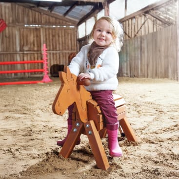 Ein kleines Kind mit hellem Haar, das einen weißen Pullover, eine kastanienbraune Hose und rosa Stiefel trägt, sitzt auf einem hölzernen Spielzeugpferd in einer sandfarbenen Scheune mit Holzwänden.