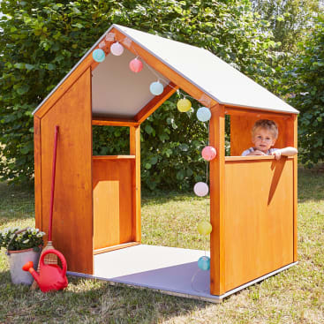 Ein kleines Kind blickt aus einem hölzernen Spielhaus mit schrägem Dach und dekorativen Lichterketten heraus. Eine rote Spielzeuggießkanne und ein Metalleimer mit Blumen stehen in der Nähe auf dem Rasen. Grüne Sträucher füllen den Hintergrund.