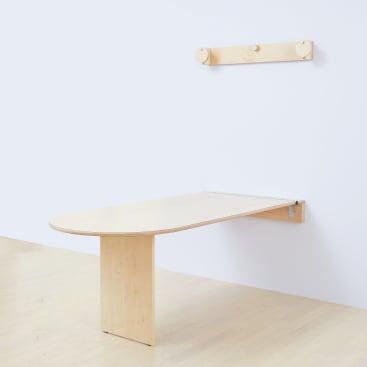 Ein minimalistischer Holztisch ist an einer weißen Wand befestigt, wobei ein Bein das andere Ende stützt. Darüber ist ein passendes hölzernes Wandregal mit drei runden Pflöcken an der Wand befestigt. Der Boden ist aus hellem Holz.