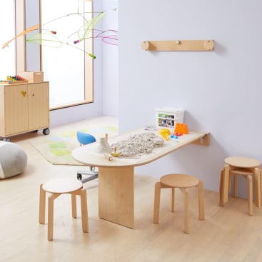 Ein Spielzimmer für Kinder mit einem Holztisch, Hockern, kinetischem Sand, Formen und Werkzeugen. Im Hintergrund befinden sich ein Aufbewahrungsschrank und ein grauer Ball, ein Mobile hängt von der Decke und Licht fällt durch ein Fenster ein.