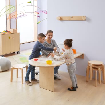 Ein Erwachsener und zwei Kinder stehen um einen hellen Holztisch in einem hellen, modernen Klassenzimmer. Die Kinder scheinen mit Knete zu spielen, und in dem Raum sind Holzmöbel und Spielzeug zu sehen.