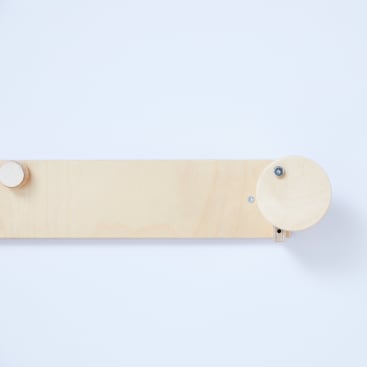Eine rechteckige Stange aus hellem Holz mit einem kleinen runden Knopf auf der linken Seite und einem größeren runden Rad auf der rechten Seite, montiert an einer schlichten weißen Wand.
