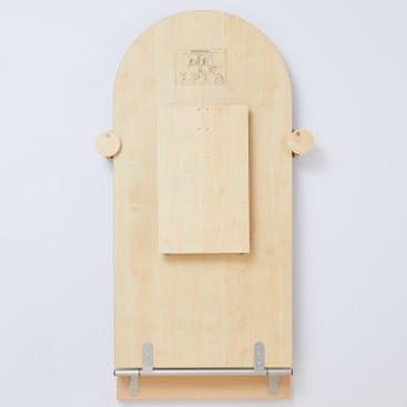 Ein rechteckiges Holzbrett mit einem kleineren Brett in der Mitte, zwei kreisförmigen seitlichen Aufsätzen, einer Metallstange am unteren Ende und eingravierten Motiven am oberen Ende, vertikal vor einem weißen Hintergrund montiert.