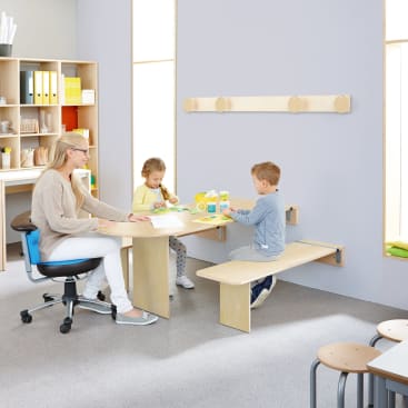 Ein Erwachsener und zwei kleine Kinder sitzen an einem Holztisch in einem Klassenzimmer. Der Erwachsene unterhält sich mit den Kindern, während sie mit Malutensilien spielen. Im Hintergrund sind Regale mit Büchern und Aufbewahrungsboxen zu sehen.