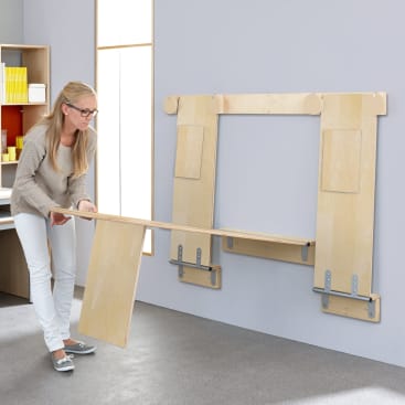 Eine Frau klappt in einem modernen Raum mit grauem Teppich und hellen Wänden einen an der Wand befestigten Holztisch mit einem Klappbein aus. Im Hintergrund sind Regale mit gelben Aufbewahrungsboxen zu sehen.