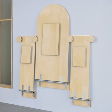 Drei unterschiedlich hohe helle Holzbretter sind vertikal an einer Wand angebracht, an denen jeweils ein rechteckiger Holzklotz mit Metallstäben in der Nähe des Bodens befestigt ist und die eine minimalistische geometrische Anordnung bilden.