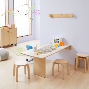 Ein Klassenzimmer mit hellen Holzmöbeln, darunter ein Tisch mit Hockern, ein Sandaktivitätsset auf dem Tisch, eine Matte, ein rollender Aufbewahrungswagen, ein Wandhaken und ein buntes hängendes Mobile. Durch ein Fenster fällt natürliches Licht ein.