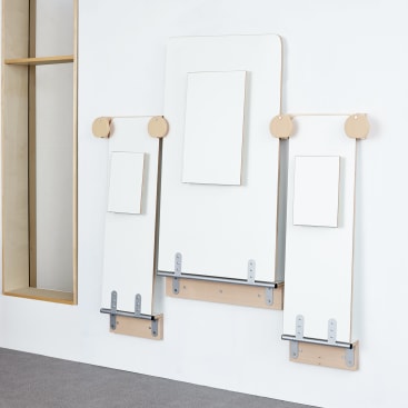 Drei weiße rechteckige Tafeln unterschiedlicher Größe sind mit Hilfe von Holz- und Metallklammern vertikal an einer weißen Wand befestigt. In der Mitte jeder Tafel ist ein kleineres weißes Rechteck angebracht. Darunter ist ein Teppichboden zu sehen.