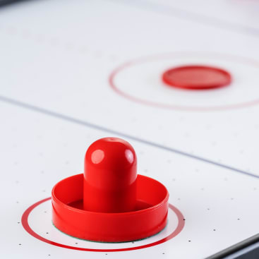 Eine Nahaufnahme eines roten Air-Hockey-Schlägers auf einem Air-Hockey-Tisch, mit einem roten Puck im Hintergrund, beide innerhalb separater roter Kreise auf der weiß gepunkteten Spielfläche.
