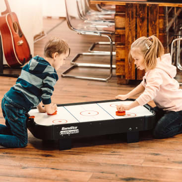 Zwei Kinder knien auf einem Holzboden und spielen ein Airhockeyspiel auf einem kleinen Tisch. Im Hintergrund sind Stühle und eine Gitarre zu sehen.