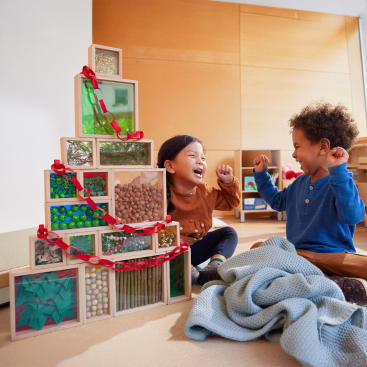 Zwei kleine Kinder sitzen in einem Haus auf dem Boden, lächeln und heben die Arme neben einer Pyramide aus transparenten Blöcken, die mit verschiedenen Gegenständen gefüllt und mit einer roten Papierkette verziert sind. Im Vordergrund ist eine blaue Decke zu sehen.