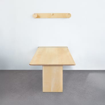 Ein minimalistischer Holztisch mit einer rechteckigen Platte und einer einzigen zentralen Stütze steht auf einem grauen Teppichboden. An einer schlichten weißen Wand hinter dem Tisch ist ein einfacher hölzerner Kleiderständer mit zwei Wäscheklammern angebracht.