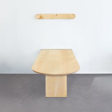 Ein heller Holztisch mit rechteckiger Basis und abgerundeten Kanten steht vor einer schlichten weißen Wand. Darüber ist ein passendes hölzernes Wandelement mit drei runden Stiften angebracht. Der Boden ist grau und strukturiert.