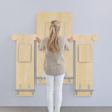 Eine Person mit langen blonden Haaren, bekleidet mit einem beigen Pullover und einer weißen Hose, steht vor einer hellen Wand mit drei montierten Holzbrettern unterschiedlicher Höhe, die jeweils mit Metallklammern und runden Holzteilen versehen sind.