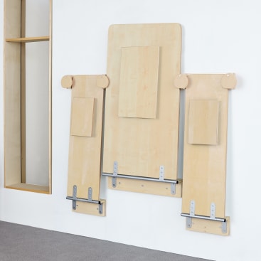 Drei helle Holzpaneele, an denen rechteckige Blöcke und Metallstangen befestigt sind, sind vertikal an einer weißen Wand angebracht. Die Paneele sind unterschiedlich hoch, und der Boden ist mit Teppich ausgelegt. Auf der linken Seite ist ein offenes Holzregal zu sehen.