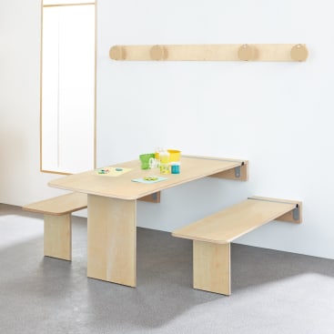 Ein Holztisch mit zwei Bänken steht vor einer weißen Wand. Auf dem Tisch stehen ein paar bunte Tassen, Teller und Blöcke. An der Wand über der Bank ist ein hölzerner Kleiderständer angebracht.