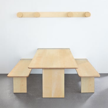 Vor einer weißen Wand steht ein heller Holztisch mit zwei daran befestigten Bänken auf beiden Seiten. Über dem Tisch ist eine Holzleiste mit vier runden Pflöcken an der Wand befestigt. Der Boden ist grau.