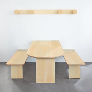 Ein minimalistischer Tisch aus hellem Holz mit zwei dazu passenden Bänken auf beiden Seiten, die vor einer schlichten weißen Wand stehen, über der eine hölzerne Garderobe angebracht ist. Der Boden ist mit einem grau gesprenkelten Teppich ausgelegt.