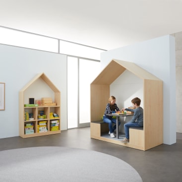 Zwei Kinder sitzen an einem kleinen Tisch in einem hölzernen Spielhaus und sind mit einer Aktivität beschäftigt. In der Nähe steht ein hausförmiges Bücherregal, das mit Büchern und Spielen gefüllt ist. Der Raum ist hell und geräumig und minimalistisch eingerichtet.