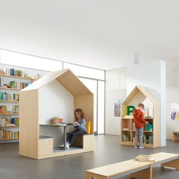 Zwei Kinder befinden sich in einer modernen Bibliothek. Eines sitzt an einem Tisch in einer hausförmigen Leseecke mit Büchern, während das andere steht und in einem Bücherregal in einer ähnlichen Ecke stöbert. Im Hintergrund stehen Regale mit Büchern.