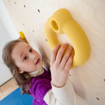Ein junges Mädchen in einem lila Hemd hält einen großen gelben Klettergriff, der an einer hellen hölzernen Kletterwand mit gleichmäßig verteilten Löchern befestigt ist.