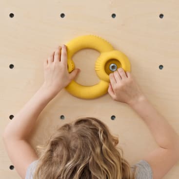 Ein Kind mit hellbraunem Haar greift mit beiden Händen nach einem gelben, runden Klettergriff, der an einer hölzernen Kletterwand mit gleichmäßig verteilten Löchern befestigt ist.