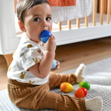 Ein Kleinkind sitzt auf einem Teppich neben einem Kinderbett und hält sich ein blaues eiförmiges Plastikspielzeug an den Mund. Mehrere bunte eiförmige Spielzeuge sind auf dem Boden neben ihnen verstreut.
