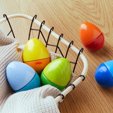 In einem mit einem beigen Tuch ausgekleideten Drahtkorb befinden sich drei eiförmige Holzspielzeuge in den Farben Blau, Gelb und Grün. In der Nähe liegen ein orangefarbenes und ein blaues Holzspielzeug in Form von Eiern auf einer Holzfläche.