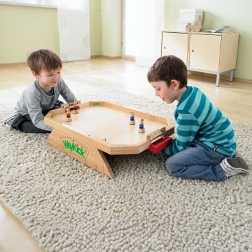 Zwei Jungen sitzen sich auf einem Teppichboden gegenüber und spielen ein Tischfußballspiel aus Holz mit kleinen Figuren und Griffen. Das Spiel ist mit "WeyKick" beschriftet. Im Hintergrund stehen ein Schrank und Spielzeug.