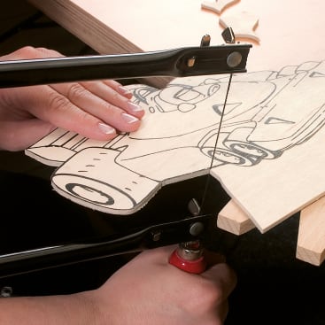 Eine Person schneidet mit einer Laubsäge ein Stück Sperrholz zu, auf dem ein Autodesign skizziert ist, und hält das Holz dabei auf einer Werkbank fest.