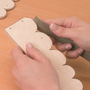 Eine Person schleift mit einem Schleifklotz die Kante eines Holzstücks mit Wellenschliff, wobei sie mit der linken Hand das Holz festhält und mit der rechten Hand schleift. Die Unterlage ist ein heller Holztisch.