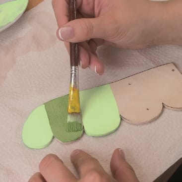 Eine Person bemalt einen Holzausschnitt mit hell- und dunkelgrüner Acrylfarbe mit einem Pinsel. Der Ausschnitt liegt auf einem Papiertuch, und nur die Hände sind sichtbar.