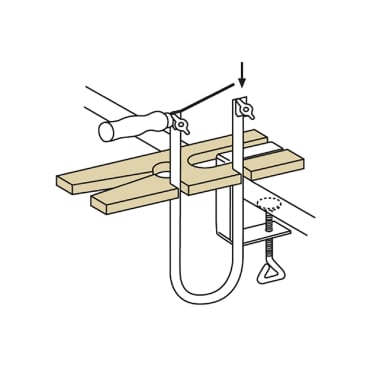 Die Abbildung zeigt eine Holzbearbeitungszwinge, mit der ein Holzbrett mit einem Loch in der Mitte auf einer Arbeitsfläche befestigt wird, während sich ein gegriffenes Werkzeug über dem Brett befindet.