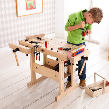Ein kleiner Junge steht an einer hölzernen Werkbank und arbeitet mit einem Schraubenzieher. Auf der Werkbank befinden sich Werkzeuge und Klemmen, und ein kleiner Werkzeugkasten steht in der Nähe auf dem Boden. Der Raum hat einen hellen Holzfußboden und eine gemusterte Tapete.