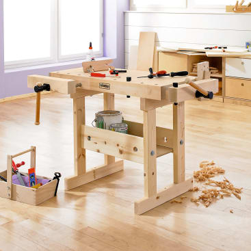 Eine hölzerne Werkbank in einer Werkstatt mit Werkzeugen, Leim und Holzspänen darauf und darum herum. In der Nähe steht ein Werkzeugkasten mit verschiedenen Handwerkzeugen auf dem Boden. Der Raum hat helle Holzböden und Regale im Hintergrund.