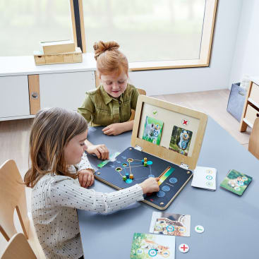 Zwei kleine Kinder sitzen an einem Tisch und spielen ein pädagogisches Brettspiel mit Tierkarten, Spielsteinen und einem hölzernen Spielbrett. Im Hintergrund sind ein großes Fenster und Regale zu sehen.