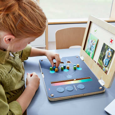 Ein rothaariges Kind spielt ein Lernspiel auf einem tablettenartigen Gerät aus Holz und ordnet farbige Stifte an. Der Bildschirm des Geräts zeigt Tierbilder und Häkchen, und das Kind sitzt an einem Tisch am Fenster.