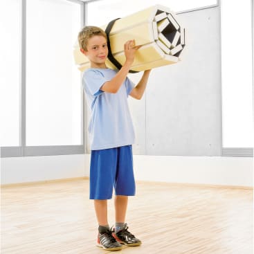 Ein kleiner Junge steht auf einem Holzboden und hält eine große geometrische Form aus Schaumstoff über seine Schulter. Er ist mit einem hellblauen T-Shirt, blauen Shorts und schwarzen Turnschuhen bekleidet. Der Hintergrund zeigt große Fenster und eine weiße Wand.