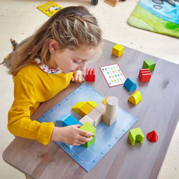Ein junges Mädchen in einem gelben Pullover sitzt an einem Tisch und ordnet bunte geometrische Holzklötze auf einer blauen Gittertafel an. Weitere Blöcke und eine Karte mit farbigen Formen und Zahlen liegen ebenfalls auf dem Tisch.