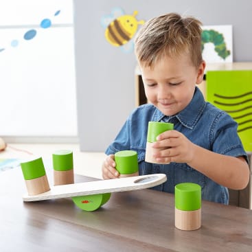 Ein kleiner Junge sitzt an einem Tisch und stapelt grüne und naturfarbene Holzteile auf einer kleinen Waage. Der Hintergrund zeigt ein dekoriertes Klassenzimmer mit einer Biene und einer Raupe an der Wand.